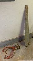 Vintage metal saw with large serrated blade leaning against a wall with one pair of red iron ice tongs beside it.