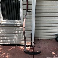 Large vintage sickle leaning against siding with long wooden handle and curved metal blade, handle paint faded and weathered.