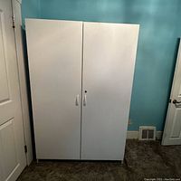 Photo of the closed white storage cabinet showing two doors and white handles against blue wall.
