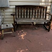 Wooden bench with backrest and armrests next to a small wooden stool on outdoor patio.