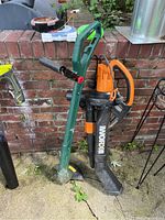 WORX corded blower and green Parkside string trimmer standing on concrete near brick wall