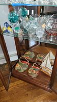 Full view of glass cabinet shelf with blown glass spheres, bowls, figurines, decorative baskets, model ship and wall hangings