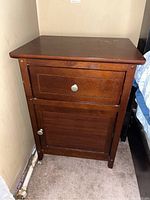 Front view of the wood nightstand showing the drawer and cabinet door with round silver knobs. The nightstand is positioned next to a wall and a bed frame is partially visible.