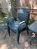 Stack of 5 green plastic framed patio chairs with woven webbing seats and backs, stacked on outdoor stone ground near metal mesh fence and glass outdoor table.