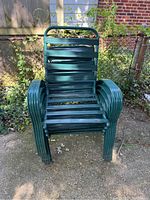 Stack of green metal slatted patio chairs shown outside on a concrete surface near foliage and fence.