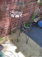 Photo of two black metal plant stands with circular glass inserts, positioned outdoors near a brick wall.