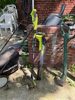 Full view of Ryobi string trimmer and leaf blower standing upright on concrete near brick wall, with some other garden items in background