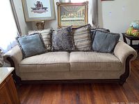 Frontal view of beige upholstered sofa with five diverse decorative pillows on a hardwood floor.
