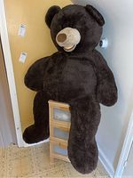Full body image of large brown stuffed bear plush leaning against a wall, showing size and shape.