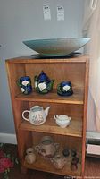 Lot overview showing wooden shelving containing the teapot, teacups, sugar bowl, sculpture vases and large glass bowl.