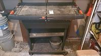Front view of the plastic tool bench cart with orange latch handles and lower shelf holding a metal bucket, on a concrete basement floor.