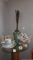 Full view of the glass table top displaying the large green vase with dried stalks, ceramic bowls and plates, fish figurines, and decorative letters spelling 'LOVE' in the background.