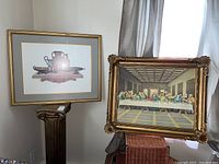 Two framed prints leaning against a wall: one Last Supper print in ornate gold frame, one classical still life with vessels in simpler gold frame