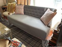 Curved grey couch with tufted backrest and solid seat cushion shown with two embroidered pink throw pillows, photographed in a living room setting.