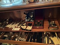 Two-tier wooden shelf showing all included flatware, cutting implements, bottle stoppers, cheese spreaders, and knife rests.