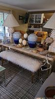 Full view of marble top sofa table with decorative items on top, including blue and white vases, table lamps with beige shades, and a long bench with white fur cushion underneath the table.