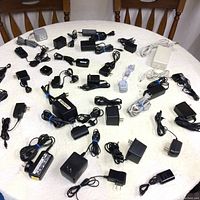 Image showing a large assortment of different types and sizes of adapters and chargers laid out on a white tablecloth, with cords tied or loosely coiled.