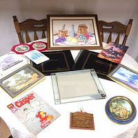 Overview of art collection items on table including Casper comic, framed paintings, multiple photo albums, copper etching, decorative plates.
