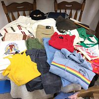 Photo showing a pile of assorted clothing items including pants, shirts, a blue hoodie with a rainbow stripe and van graphic, a yellow tank top, a red top, and a black beret.