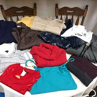Overview of all clothing items laid on chairs and table including blouses, sweaters, jackets, and blazers in multiple colors and styles.