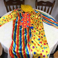 Full view of clown jumpsuit laid flat showing vertical rainbow stripes on one half and yellow with colorful polka dots on the other, with attached red tie with circular patterns.