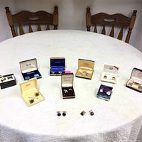 Photo showing assortment of cuff links and tie clips displayed on a round white table with wooden chairs in background, multiple pairs in varied original boxes and loose