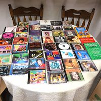 Image showing 44 CDs and several entertainment/trivia books on a white tablecloth with wooden chairs in background. CDs spread out in rows, covers visible with artists such as Cher and Elton John.