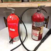 Photo of two red fire extinguishers on a table, one with hose and nozzle, the other missing nozzle.