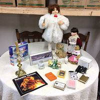 Overall view of vintage dolls, plush bears, porcelain figurines, brass candle holder, framed photo, playing cards, and other miscellaneous items on round table covered with white tablecloth.