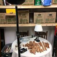 View of halogen lamp, small table lamp, glass containers, and wooden wine rack parts on a round table with stacked U-Haul boxes in the background.