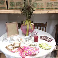 Overall view of various household decorative items including vases, picture frames, porcelain figurines, candle holders, apron and a candle arranged on a round white table.