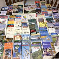 Overhead photo showing a large assortment of vintage maps and travel guides spread out on a wooden table, featuring titles for New York, Nova Scotia, Prince Edward Island, Edmunston, Windsor, East Texas, and more.