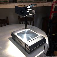 Full view of the overhead projector on a table, showing the projection surface and the arm with mirror/lens assembly.