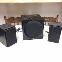 Front view showing two satellite speakers flanking a larger subwoofer unit on a white surface with wooden chairs behind.