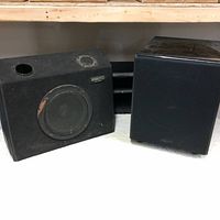 Two speakers placed side by side on a shelf, Kenwood branded on the left speaker, the right one has a cloth cover.