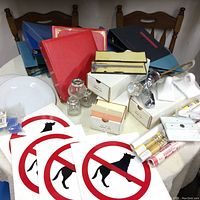Overview of lot items on table showing No Dogs signs, binders, glass spice jars, bathroom tap in box, small glass bottles, and office supplies.
