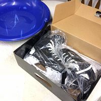 Pair of black and white Hxi hockey skates packed in a shoebox with plastic wrapping, placed beside blue flying saucer sled.