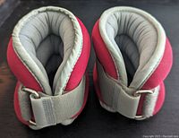 Pair of ankle weights with red outer fabric and grey padded interior, showing adjustable strap with metal D-ring.