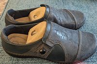 Close-up of a pair of black Clarke's slip-on shoes with side zipper, showing textured leather finish and shoe form inside.