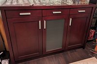 Mahogany sideboard showing three drawers on top and three cupboards below, one cupboard with frosted glass door, all with silver handles, on a dark floor.