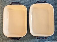 Top-down view showing two rectangular casserole dishes with cream interior and blue handles embossed with STAUB