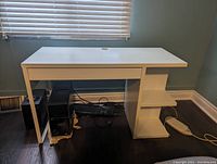 Front view of white computer desk showing single drawer on left, two open rectangular shelves on right under the desk surface, and two open cube shelves at the bottom right.
