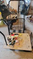 Photo showing a black metal grocery buggy with foldable wire basket, a wooden flat utility cart with black handle, and multiple rolls of tape placed on the wooden cart surface, along with small miscellaneous hardware items.