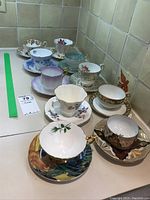 Overall assortment of 10 tea cups and 10 matching saucers placed on a countertop with various patterns and colors, includes one chipped saucer.