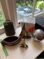 Photo shows an 8 inch rustic brass cow bell, two brass sailboat figurines, a metal magnetic globe puzzle on a clear stand, and a ceramic pot shaped like a fish with decorative painting.
