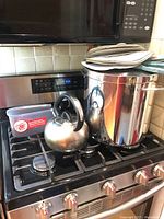 Stainless steel lobster pot, kettle, plastic container with SoftScrape label and stacked cloth serviettes on a gas stove.