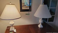 Pair of white marble table lamps with twisted design bodies and square bases, topped with white shades, positioned on wooden table.