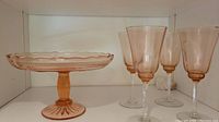 Pink glass dessert tray and four pink stemmed glasses displayed on shelf, one glass has a chip.
