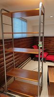 Front angle view of metal frame shelving unit with five brown wood shelves, positioned in a room with wooden paneling.