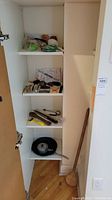 Cabinet shelf with various kitchen utensils including knives, peelers, small pan, and large wooden spoon leaning against the wall